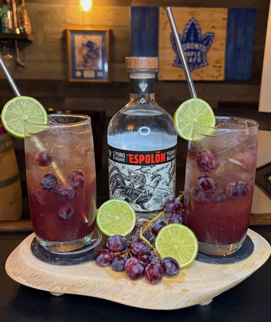 The Tequila Transfusion cocktail recipe by Millers Amish Pub, featuring two purple grape and ginger ale cocktails in tall glasses next to a bottle of Espolòn Blanco Tequila, fresh concord grapes, and lime wheels.