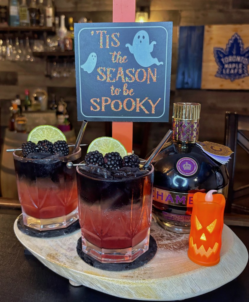 Two layered Halloween cocktails with dark purple and red gradients, garnished with blackberries and lime wheels on a wooden tray next to a bottle of Chambord liqueur and a glowing orange jack-o'-lantern candle.