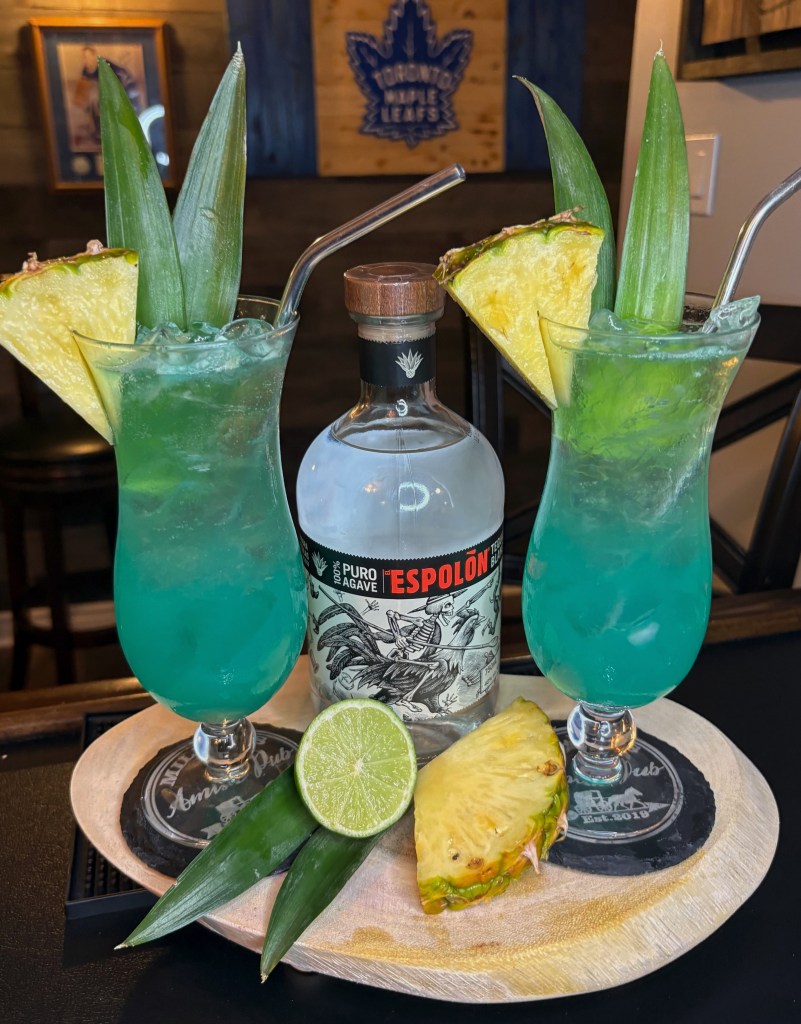 Two vibrant electric blue cocktails garnished with pineapple slices and leaves, served on a wooden tray with a bottle of Espolón Tequila, a fresh lime half, and a pineapple wedge at Miller’s Amish Pub.