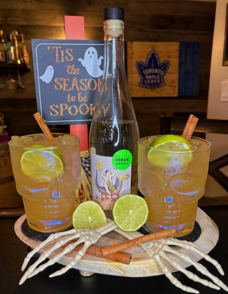 Two Mez-Skull Mule cocktails in skull glasses garnished with lime and cinnamon sticks, arranged on a Halloween display tray with a bottle of Mezcal Verde Amaras, skeleton hands, and a spooky sign.