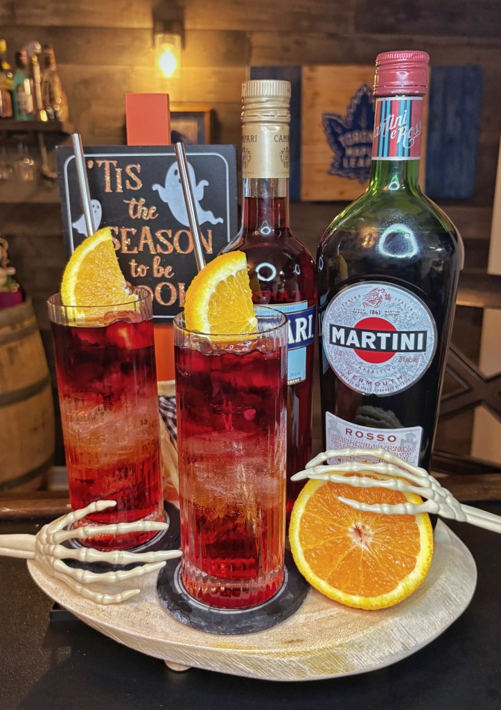 Two Halloween-themed red Americano cocktails in tall fluted glasses garnished with orange slices on a wooden tray alongside bottles of Campari and Martini Rosso sweet vermouth.