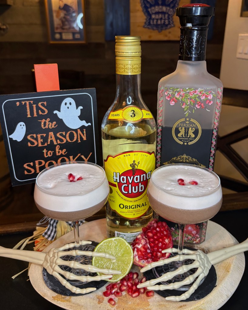 Two foaming pink pomegranate rum sour cocktails in coupe glasses sitting on a rustic wooden tray. The glasses are held by decorative white skeleton hands. In the background are bottles of Havana Club 3 Year Rum and Persian Empire Pomegranate Liqueur, alongside a chalkboard sign that reads "'Tis the season to be spooky" next to a fresh split pomegranate.