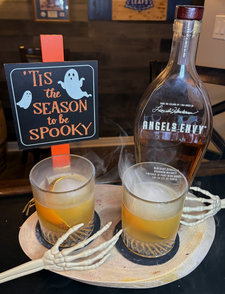 "Two smoked bourbon Old Fashioned cocktails served with ice spheres and orange twists on a tray with plastic skeleton hands, next to an Angel's Envy whiskey bottle and a 'Tis the Season to be Spooky' sign."